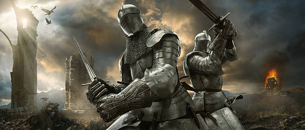 Two Medieval Knights With Swords On Battlefield Near Ruined Monuments A dramatic image of two medieval knights in full suits of armour and chainmail holding swords in fighting positions close to stone monument ruins and burning castle in the background. The knights are on a high battle ground under a dramatic cloudy evening sky with shafts of sunlight and ravens. battle photos stock pictures, royalty-free photos & images