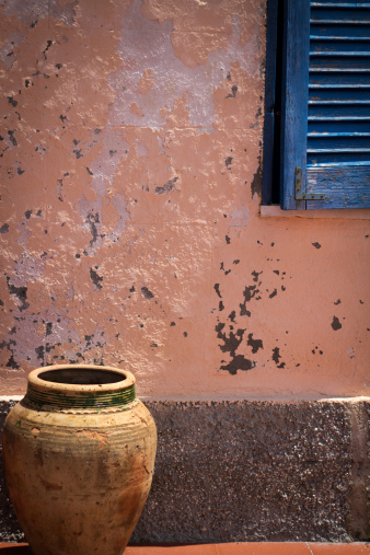 Aeolian Islands still life: old ceramic olive jar, mottled pink wall, and blue shutters. A brightly lit scene with colors typical of the island. The Aeolian Islands are part of Sicily, Italy, and located just north of Messina. The pot is a traditional olive oil container.