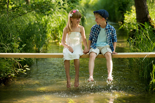Small girl and boy looking at each other, holding hands while sitting on wood over the stream in forest.