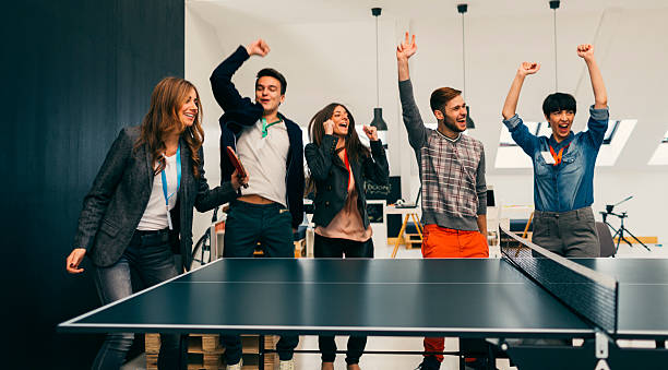 Young Business People Playing Table Tennis In Their Office. Group of young business people running new start up company. They have table tennis in their office. Two colleague playing table tennis and other coworkers cheering. Jumping and smiling. Location is released. office competition stock pictures, royalty-free photos & images