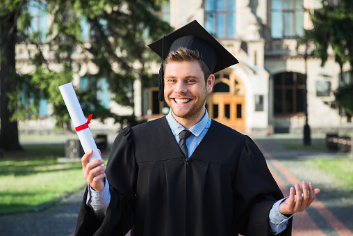 Young male student dressed in black graduation gown. Campus as a background. Boy cheerfully smiling, holding diploma and looking at camera
