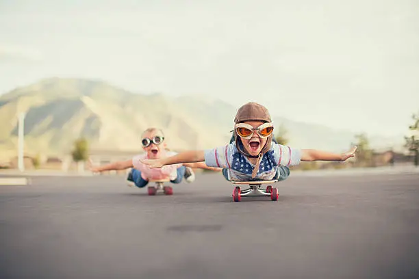 A young boy and girl are wearing flying goggles while outstretching their arms to attempt flying while on skateboards. They have large smiles and are imagining taking off into the sky.