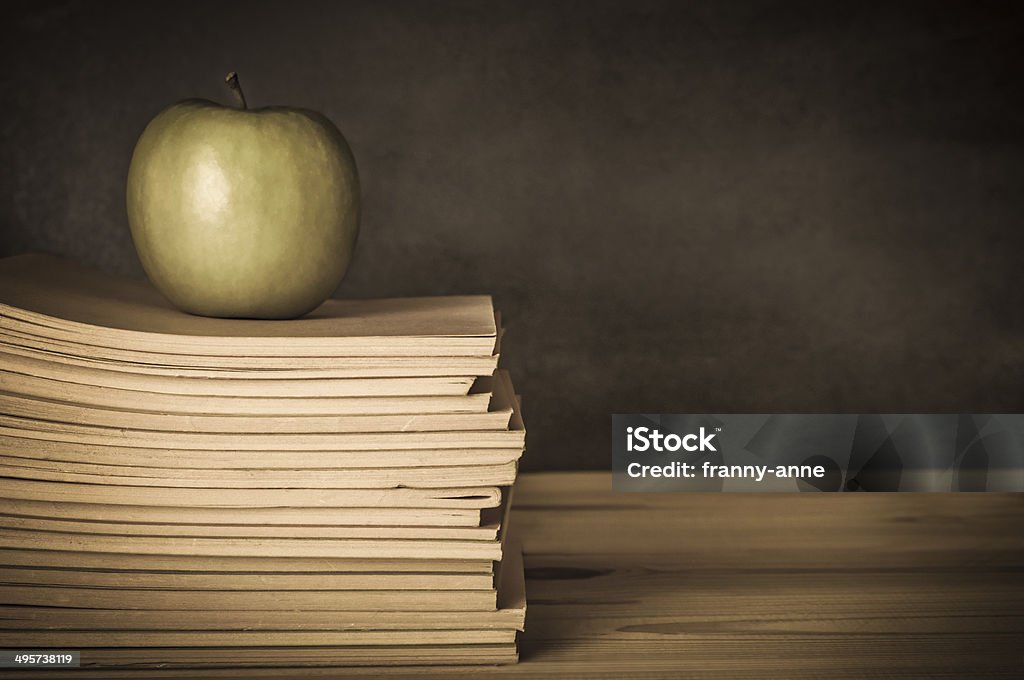 Teacher's Desk - Apple on Books Vintage style image of a teacher's wooden desk, with an apple on top of a pile of used, worn, exercise books.  Blackboard in soft focus background.  Undersaturated hues. Desk Stock Photo