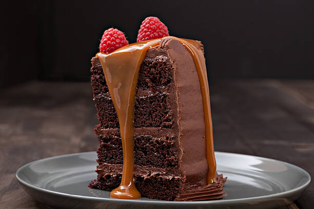 Slice Of Chocolate Fudge Cake An extreme close up horizontal photograph of a generous slice of chocolate fudge cake toped with two fresh raspberries and with caramel sauce flowing down the side and frosted back. Isolated on black. chocolate cake stock pictures, royalty-free photos & images