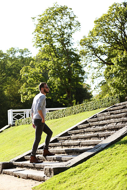 Man wearing jeans,boots and a grey shirt walking up a stone steps outside on a sunny summer day. 