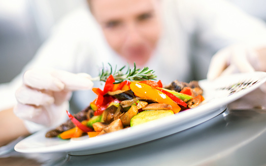 Blurry mid-aged female chef seasoning a salmon steak meal with rosemary sprig.Wearing white uniform and white protective gloves when touching ready to eat food. Prepared meals are in the background