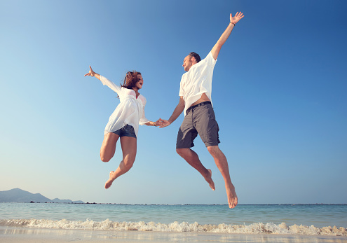 Playful couple having fun during summer while jumping together on the beach.