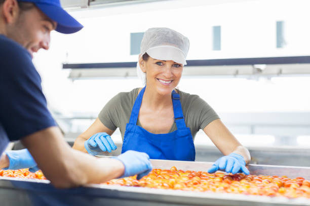A school cook wearing a white uniform, apron, and hairnet smiles at the camera while working in the cafeteria during lunchtime. The background shows part of a typical Brazilian school environment, with students in uniforms and green-painted walls. This image highlights the essential role of school cafeteria staff in student nutrition and the daily routine of public education