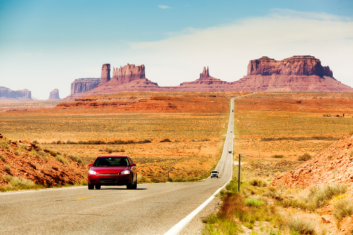 Subject: Tourists in a red car traveling in the American Southwest, driving downhill on a straight length of highway stretching from southern Utah toward Monument Valley, Arizona