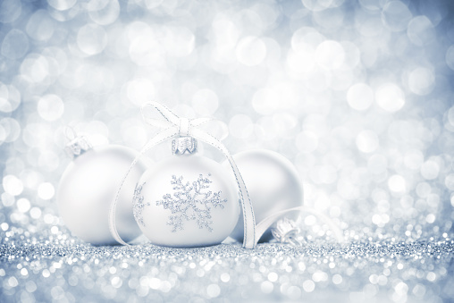 Three silver bauble with snowflake decoration and ribbon on glitter. Cold gray vintage glitter defocused lights abstract background. Light centre and corner vignette. Shallow depth of field. Light purple tint.