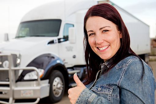 A royalty free image from the trucking and transportation industry of a female truck driver giving a postive thumbs up hand signal while smiling and standing in front of her white semi truck cargo container.