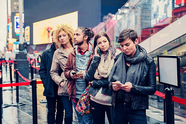 Young people waiting in line to buy tickets in NewYork. Young people waiting in line to buy tickets in New York. While waiting they are on the phone or texting. Blank placard on the right. Focus on the girl with short hair. It's raining lightly and they are in Times Square. Waist up outdoors horizontal shot. people in a line stock pictures, royalty-free photos & images