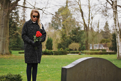 Young woman standing at graveside of a deceased family member. Female at cemetery grieving holding flowers.