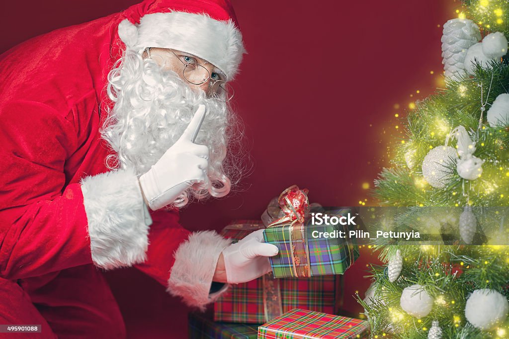 Santa Claus putting gift box or present under Christmas tree Santa Claus putting gift box or present under Christmas tree at eve night. It's a secret. Surprise! Don't tell the kids! Xmas and New Year holiday! Copy space at red background 2015 Stock Photo