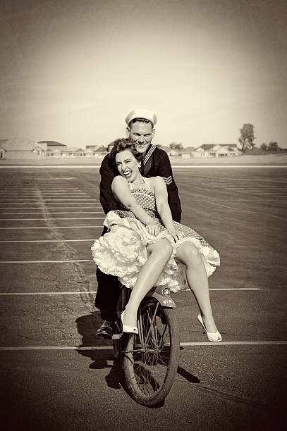 WWII Navy Man and His Pretty Woman Riding A Bike WWII Navy Man and His Pretty Woman Riding A Bike. They are laughing and having fun as she tries to keep her dress from blowing up in the wind.  Warm, happy and iconic.  Some Grain. navy photos stock pictures, royalty-free photos & images