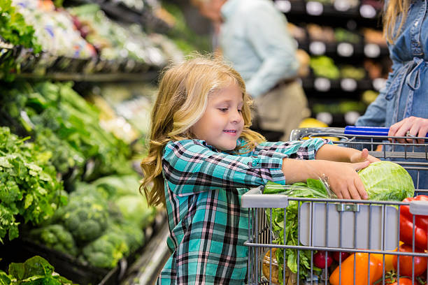 Little girl helping mother shop for produce in grocery store Elementary age Caucasian blonde little girl is smiling while placing lettuce in shopping cart. Child is shopping for produce and healthy food with her mother in local grocery store. Child is wearing a plaid shirt. Other customers are shopping in background. farmers market healthy lifestyle choice people stock pictures, royalty-free photos & images