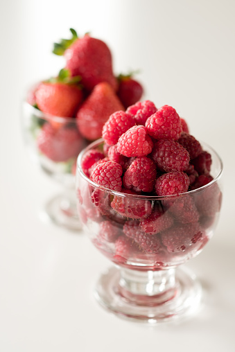 Glass cup of raspberries and cup of starberries on white table.