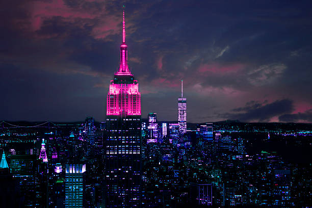 Aerial view of Manhattan, New York City, USA skyline at night.  The Empire State building is lit in pink at the top, foreground.  The rest of the cityscape is lit in lavender hues as the dramatic twilight sky looms overhead.  The Freedom Tower (One World Trade Center) can be seen in the far distance.  Manhattan Bridge in background left. 