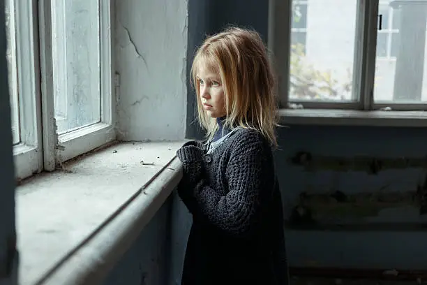Hopeless life. Close up of depressed poor little girl standing near window and looking aside while feeling miserable