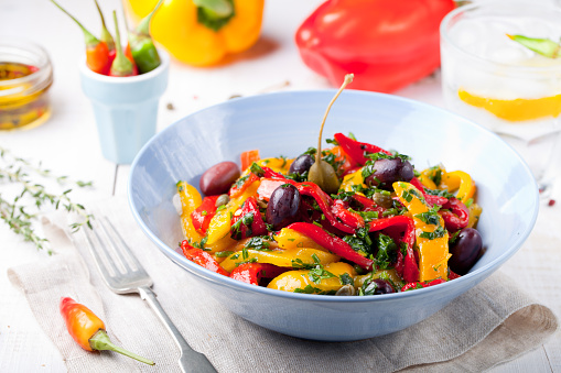 Roasted yellow and red bell pepper salad with capers and olives in a blue bowl on a white background. Grilled vegetables.