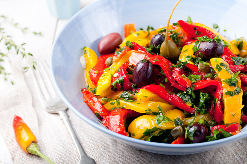Roasted yellow and red bell pepper salad with capers and olives in a blue bowl on a white background. Grilled vegetables.
