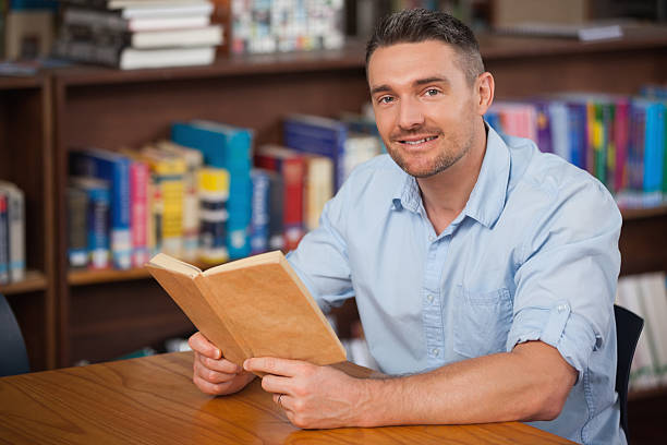 istock Smiling male student reading book in college library 496157957
