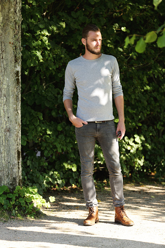 An attractive man wearing jeans,boots and a shirt standing with a green background on a sunny summer day.