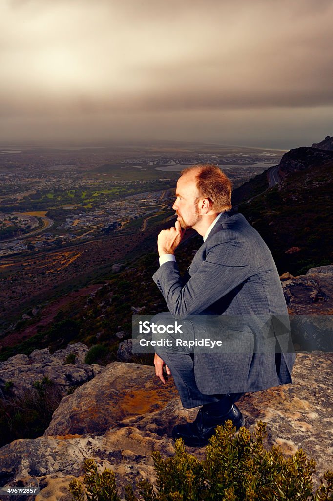 What does the future hold? Worried businessman considering his prospects A businessman crouches on a hilltop, his hand to his chin, looking worriedly out over a dark, moody cityscape, possibly considering his future prospects. Copy space on the clouded sky. 50-59 Years Stock Photo