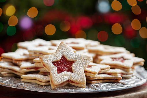 Plate of raspberry jam sandwich sugar Christmas cookies in star shaped cutout under the Christmas tree with defocused lights