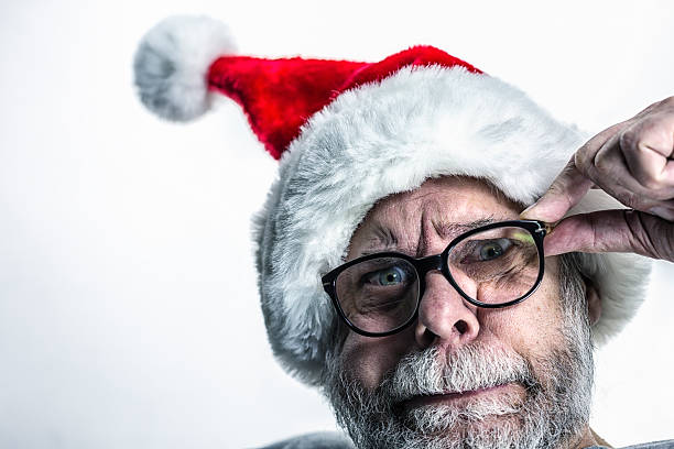 A bizarre, nerdy senior adult man wearing a Santa Claus hat is staring fearfully at the camera as he adjusts his black thick rimmed bifocal eyeglasses.