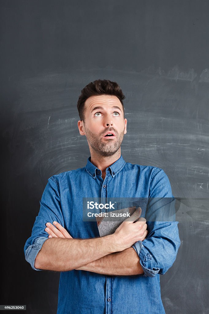 Surprised man against blackboard, holding a book in hands Studio portrait of surprised adult man wearing jeans shirt, standing against blackboard, looking up with mouth open. Men Stock Photo