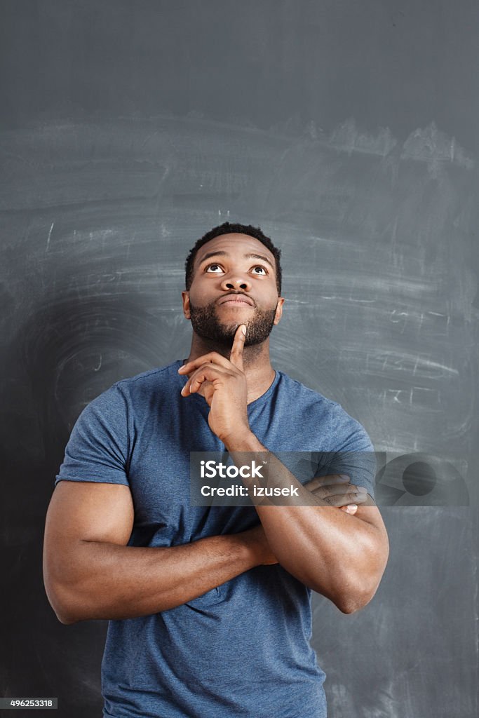 Pensive afro american man against blackboard Studio portrait of pensive afro american man, standing against blackboard, looking up with finger on chin. Asking Stock Photo