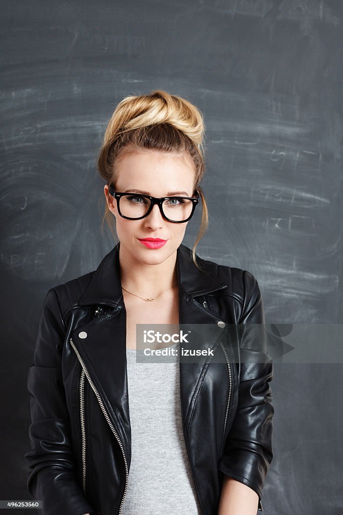 Blonde young woman against blackboard Studio portrait of beautiful blonde woman wearing leather jacket and nred glasses, standing against blackboard, looking at camera. Women Stock Photo