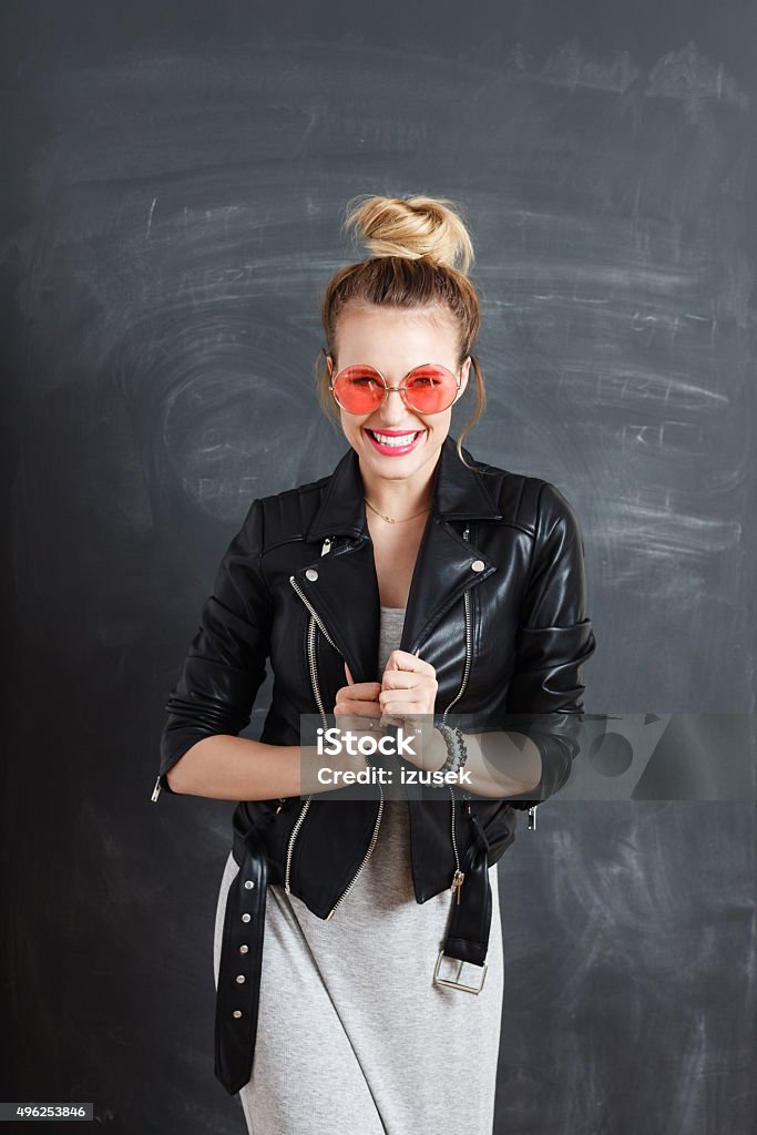 Happy young woman against blackboard Studio portrait of beautiful blonde woman wearing leather jacket and pink glasses, standing against blackboard, laughing at camera. Ecstatic Stock Photo
