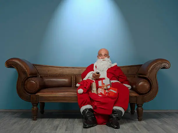Looser lonely Saint Nicholas sitting on sofa in dark and watching television.He looks quite tired and unhappy.There are gift boxes in bag by his leg.Background is blue.Shot in horizontal framing with medium format camera Hasselblad