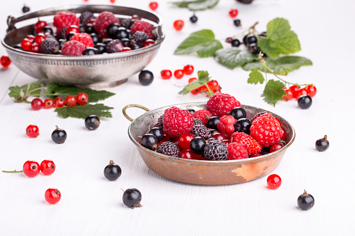 Ripe berries in a metal bowl on a white wooden background. Currants, raspberries, blackberries and wild strawberries.