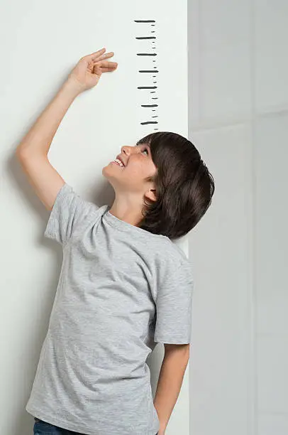 Closeup of little boy measuring height himself against white wall. Smiling cute boy measures his height. Boy growing tall. Young boy checking his height with the hand.
