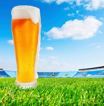 Ice Cold  Glass of  Beer , covered with water drops - condensation. Glass standing on  perfect grass, football stadium in the background.