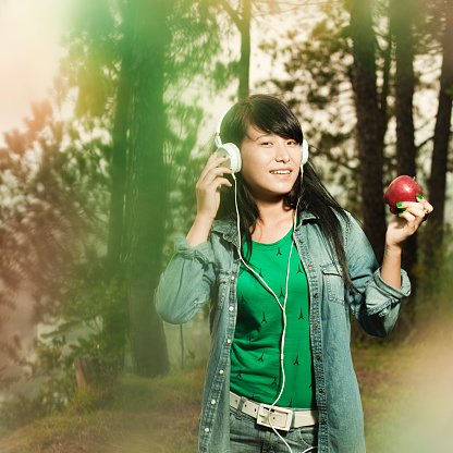Close-up, outdoor image shot through blur objects by using selective focus of a serene happy Asian teenage girl listening music through headphones, standing in beautiful nature full of plants and trees. She is in blue denim shirt, green top and jeans. Square composition with copy space and selective focus.