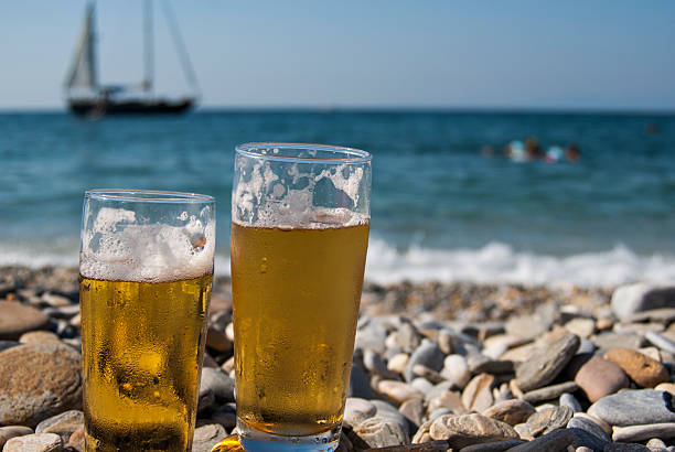 A glass of the cold beer on the table disposed on the outdoor patio, seascape and the cloudy sky are in the background