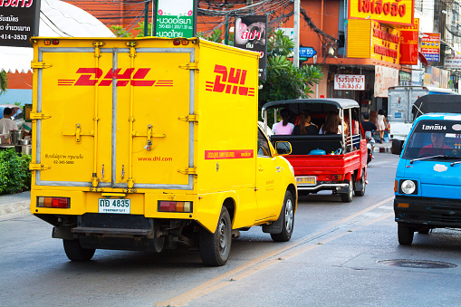 Bangkok, Thailand - April 30, 2014: Pick-up truck of German Deutsche Post and DHL is driving in street Chokchai 4 in Ladprao. Behind car people are on sidewalk. In street are tuk-tuks driving and transporting passengers in both ways. At right side driver is sitting alone in front seats of tuk-tuk.