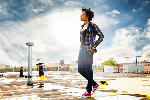 Confident, hip, young black woman walking on wet rooftop back lit by the sun, hands in back pockets. She is wearing jeans, a plaid shirt, sneakers and sporting a short afro.
