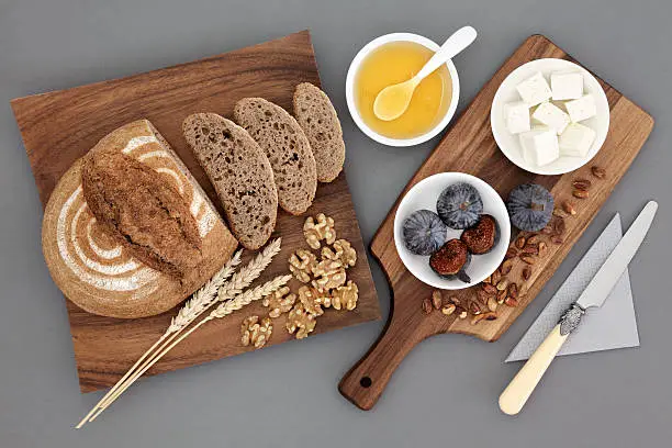 Greek snack food with feta cheese, honey, figs, walnuts and pistachio nuts with rye bread and wheat sheaths on maple wood board over grey background.