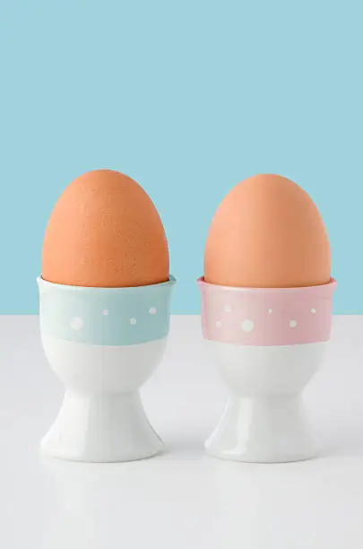 Two egg cups, decorated with pink and blue stripes with white spots and holding brown eggs on white table with light blue background.  Copy space above.