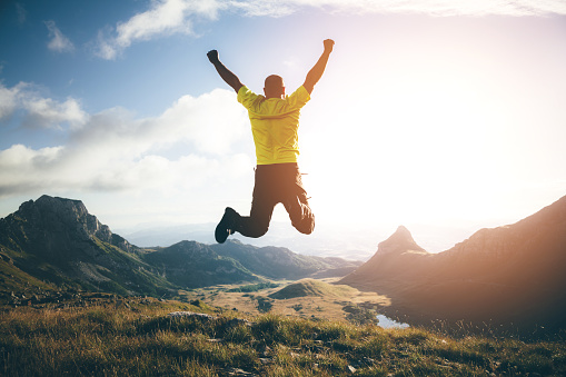 Success, achievement concept. Man jumping in the air on top of the mountain, hands raised up, mountain landscape on background.