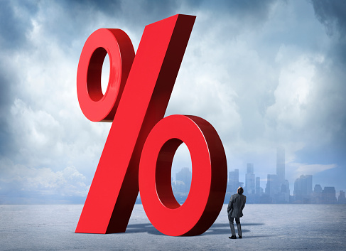 A businessman looking up at a red percent sign representing rising interest rates. There is a city skyline in the background with stormy clouds above.
