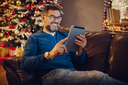 Mature man sitting on sofa in front of Christmas tree, using digital tablet and checking news, sport scores or planning vacation.Evening or night with beautiful yellow lights lightning the scenes.