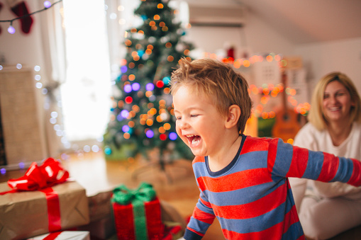 Little boy is running thrilled after he got the Christmas presents he wanted on Christmas morning. Both mother and son are wearing pyjamas, since they are sitting in their lovely decorated living room. 