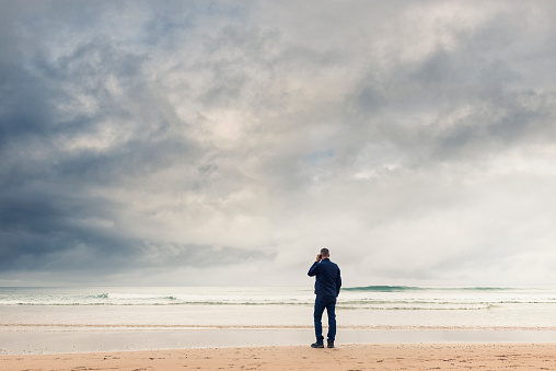 man stood looking out to sea using mobile phone, wearing a jacket on a cloudy moody Autumn beach. North Cornwall.