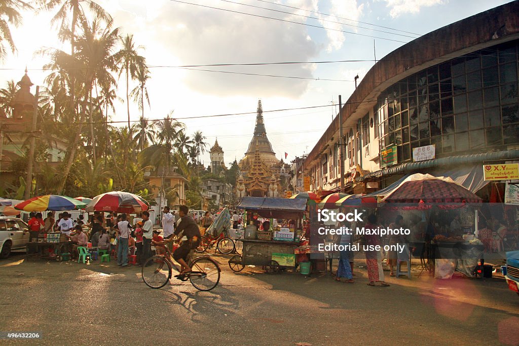 People crowding a street market in Yangon, Myanmar Yangon, Myanmar - January 4, 2009: Crowd of people walking in a street market of Yangon, Myanmar. Some guys stand by the stalls, while other walk and ride bicycle. The Shwedagon Pagoda, the main city temple, is visible in the background. Warm sunlight illuminates the scene through palm trees. Yangon Stock Photo
